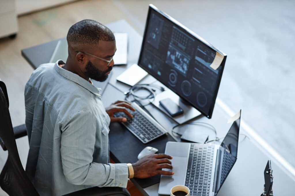 A data analyst working on multiple screens displaying data visualizations and analytics, using a laptop and desktop monitor in a modern office setup.