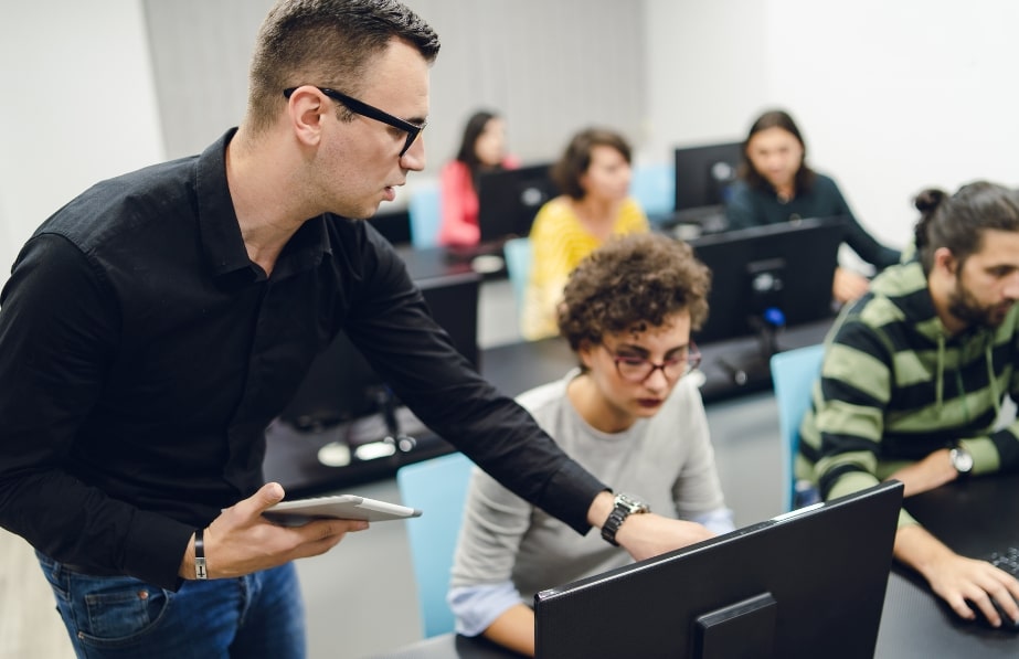 A teacher assisting a student in a computer lab classroom, with several other students working on desktop computers in the background.