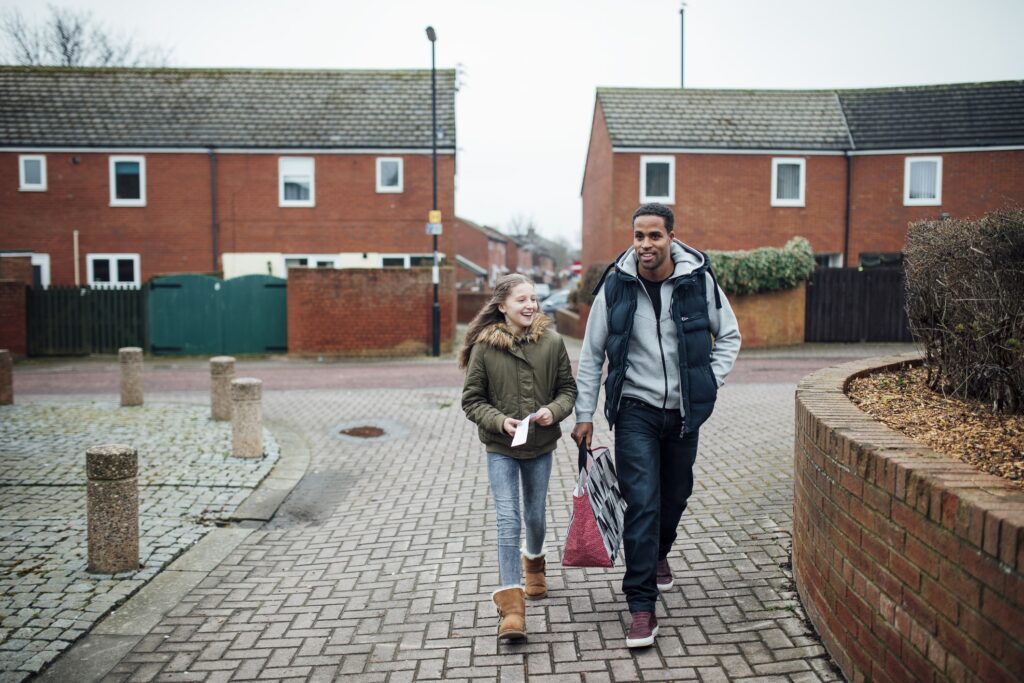 A man and a young girl walking through a residential neighborhood with red-brick houses, both smiling and holding shopping bags on a cloudy day.