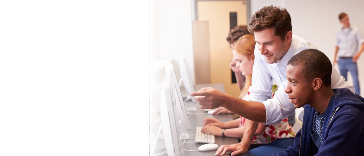 A teacher assisting a student at a computer, while other students work on computers in a classroom setting, with two people talking in the background.