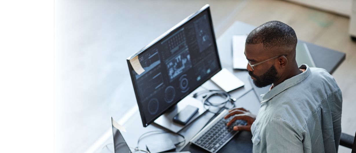 A data analyst working on multiple screens displaying data visualizations and analytics, using a laptop and desktop monitor in a modern office setup.