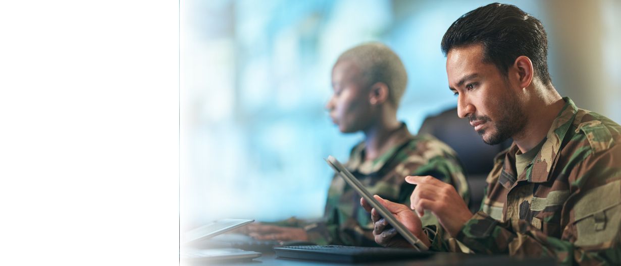 Two military personnel in camouflage uniforms working on digital devices in a technology-focused command center.