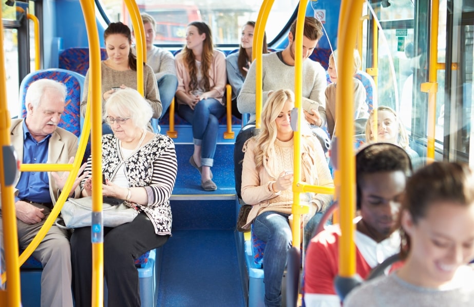 Passengers of various ages sitting on a public bus, some using mobile phones, while others are engaged in conversation during daylight.