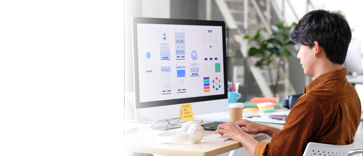 Person designing a user interface on a computer, with wireframes and flowcharts on the screen, alongside headphones and a coffee cup on the desk.