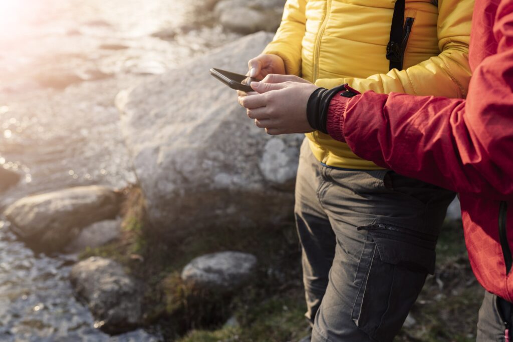 Two people stand by a river, closely examining a smartphone in their hands as they engage in conversation.