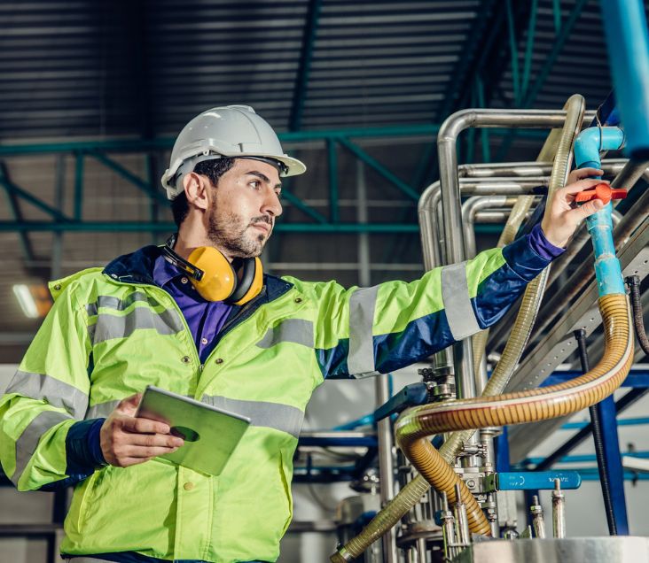 A worker in a high-visibility jacket and hard hat uses a tablet while inspecting a machine in a factory or industrial setting.