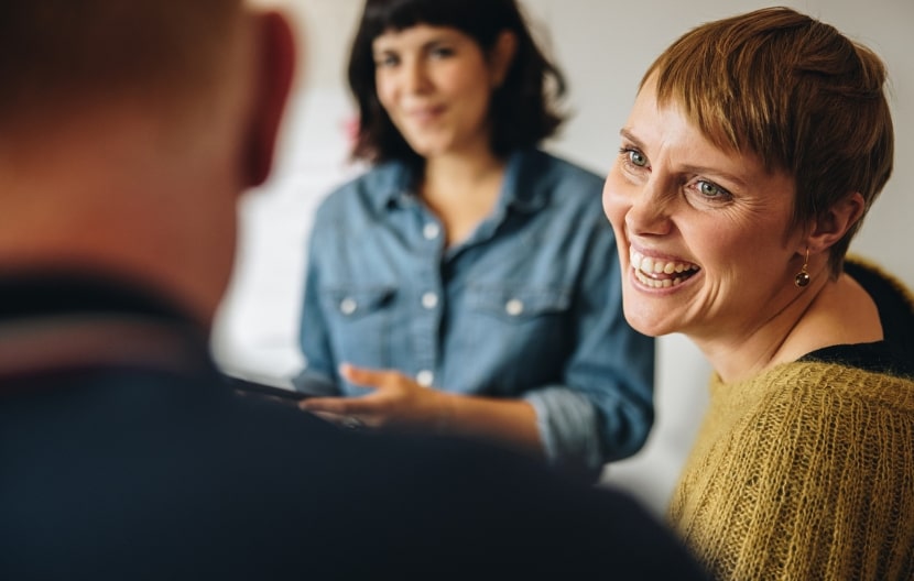 A smiling woman in a knitted sweater engages with another woman in a denim shirt, creating a warm, collaborative atmosphere in a professional setting.