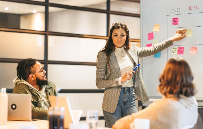 A woman leading a meeting by explaining tasks on a whiteboard with sticky notes, while two colleagues listen and discuss in a modern office setting.