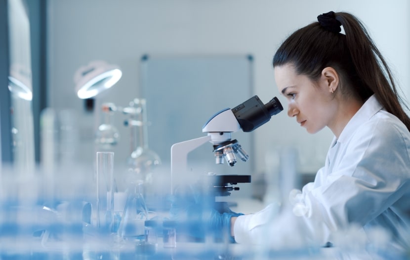 A female scientist wearing a lab coat using a microscope in a laboratory, surrounded by lab equipment and glassware.