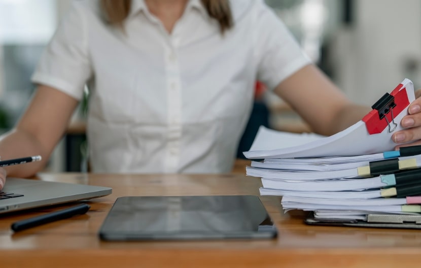 A woman working at a desk with a laptop, organizing a large stack of documents clipped together, in an office environment.