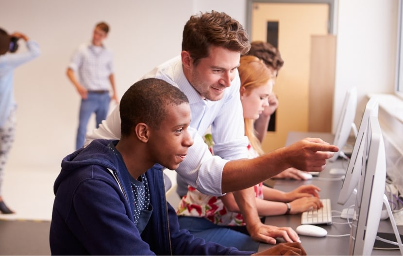 A teacher assisting a student at a computer, while other students work on computers in a classroom setting, with two people talking in the background.