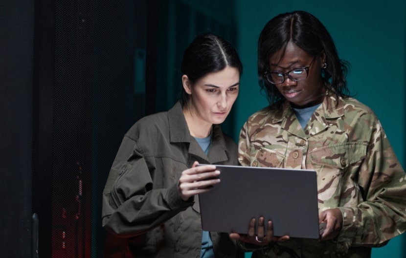 Two women in military or tactical attire are working together, reviewing data on a laptop in a server room or data center environment.