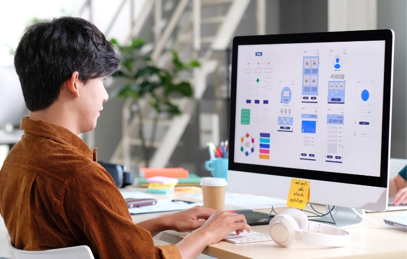 Person designing a user interface on a computer, with wireframes and flowcharts on the screen, alongside headphones and a coffee cup on the desk.