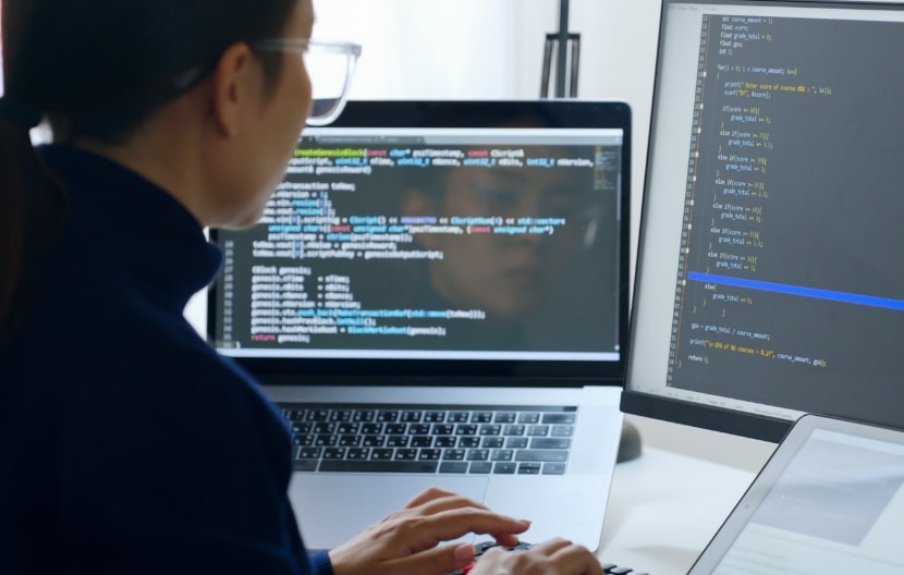 A software engineer working on a laptop and desktop monitor, writing and debugging code, with multiple screens displaying programming code in a modern workspace.