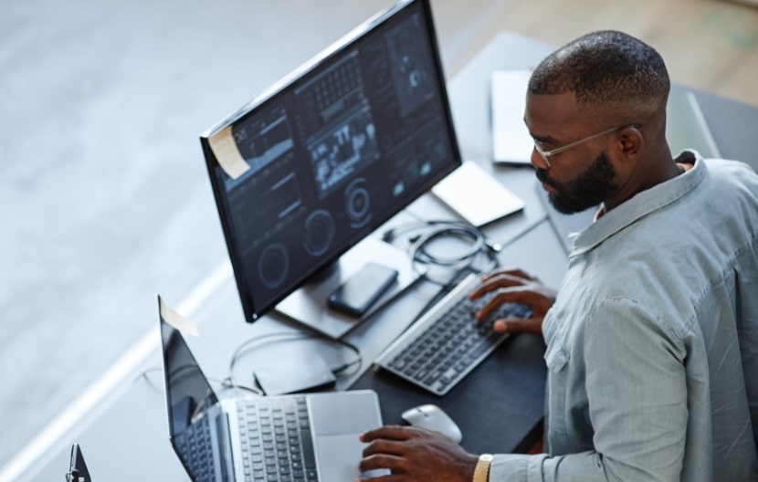 A data analyst working on multiple screens displaying data visualizations and analytics, using a laptop and desktop monitor in a modern office setup.