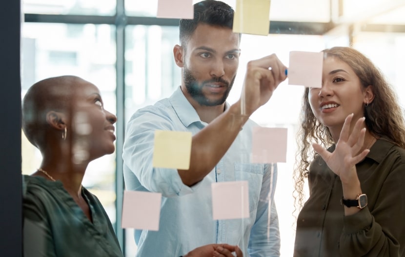 A diverse team of professionals brainstorming together by placing sticky notes on a glass wall in a modern office.