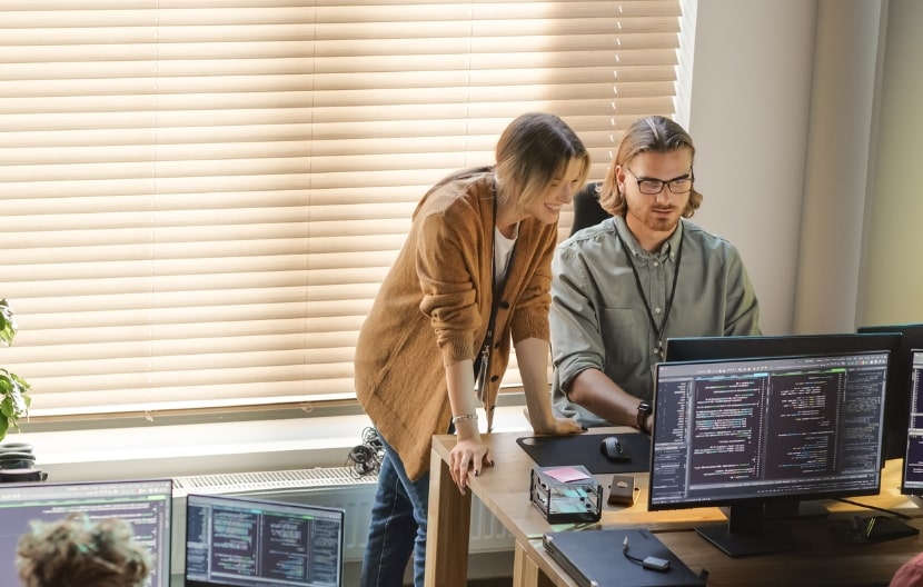 Team of developers working at desks with multiple monitors displaying code, collaborating in a modern office setting.